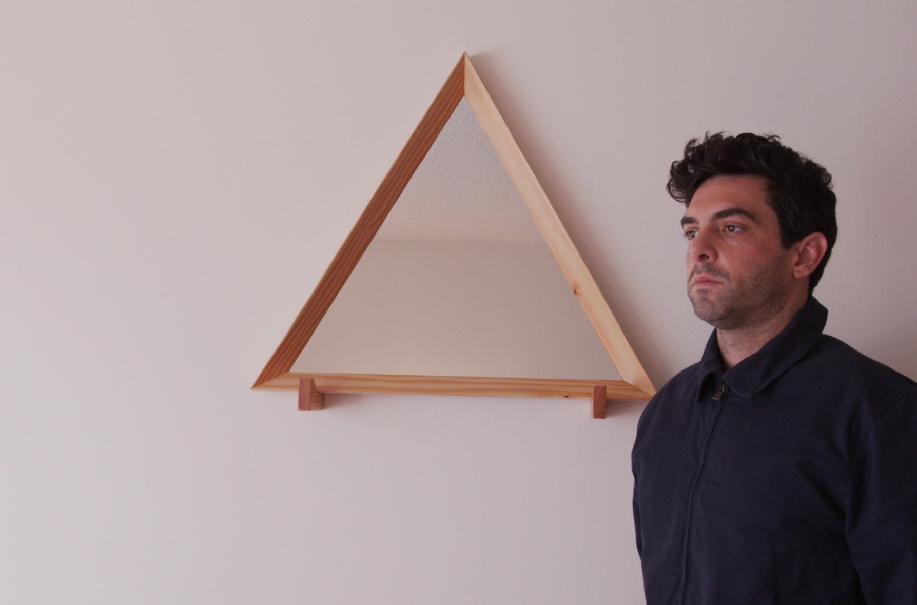 Triangle-shaped wooden mirror mounted on a wall with a natural wood frame, accompanied by a person standing beside it. The mirror adds a minimalist and geometric touch to the room decor.
